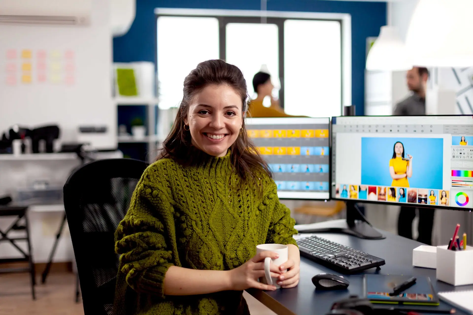 Woman retoucher looking at camera smiling sitting in creative media agency retouching client photos on PC with two displays. Photographer graphic editor working in start up office with digital assets